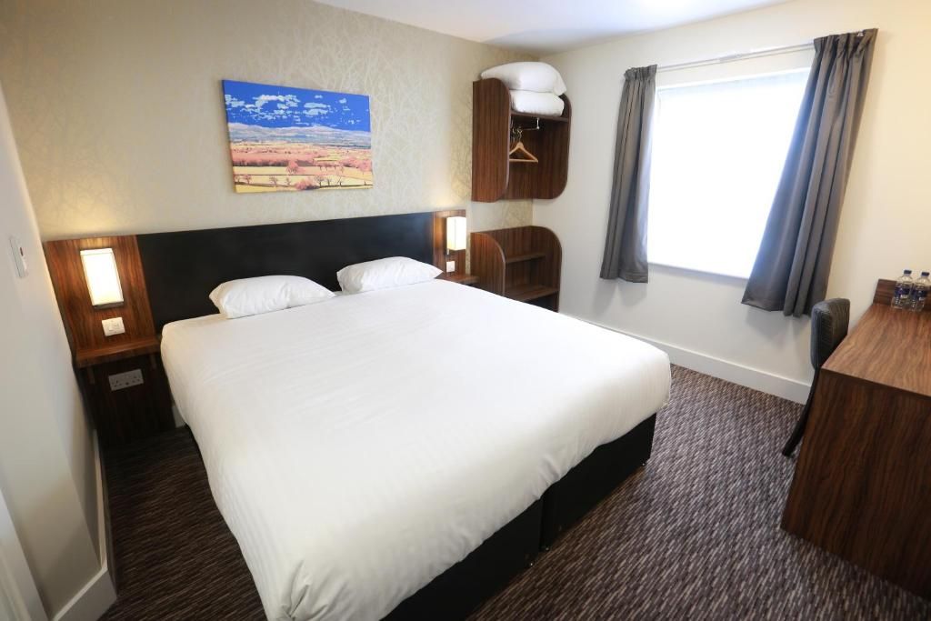 A hotel room with a large bed, a nightstand, a wooden wardrobe, a desk, and a window with curtains.