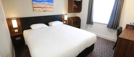 A hotel room with a large bed, a nightstand, a wooden wardrobe, a desk, and a window with curtains.