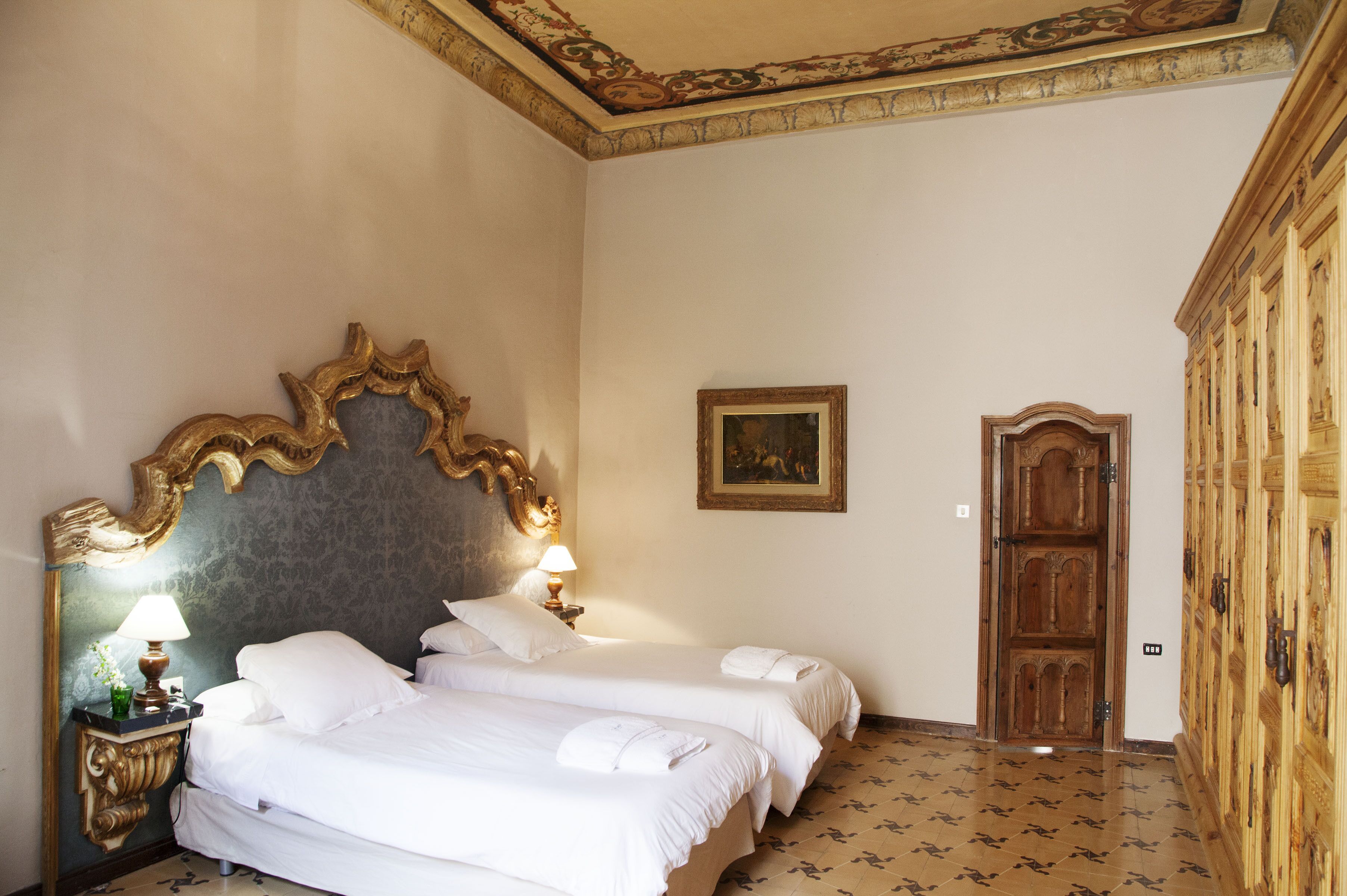 Habitación de hotel con dos camas, un espejo grande y ornamentado, un armario de madera y un cuadro enmarcado en la pared.