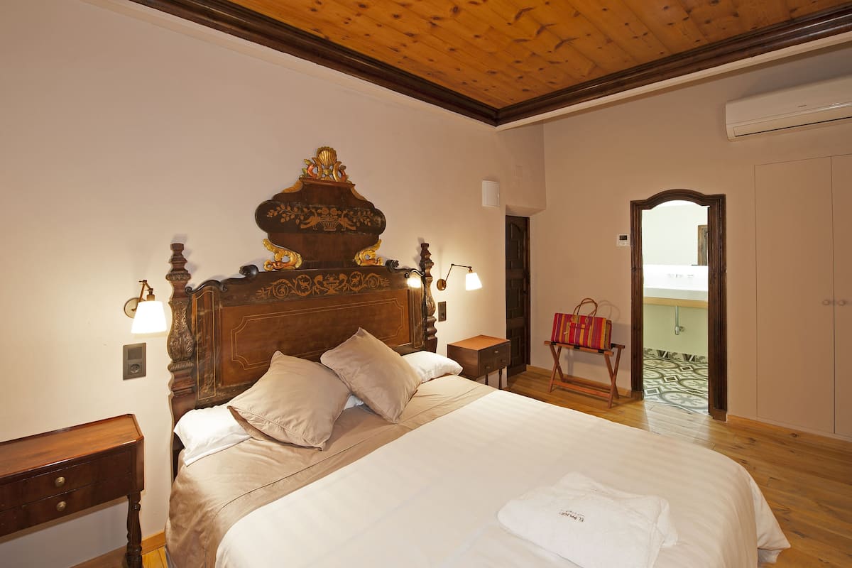 Une chambre à coucher avec un grand lit, une tête de lit en bois, des lampes de chevet et une vue sur l’extérieur par une porte ouverte.