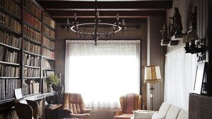Een gezellige bibliotheek met lederen fauteuils, een kroonluchter en een groot raam met transparante gordijnen.