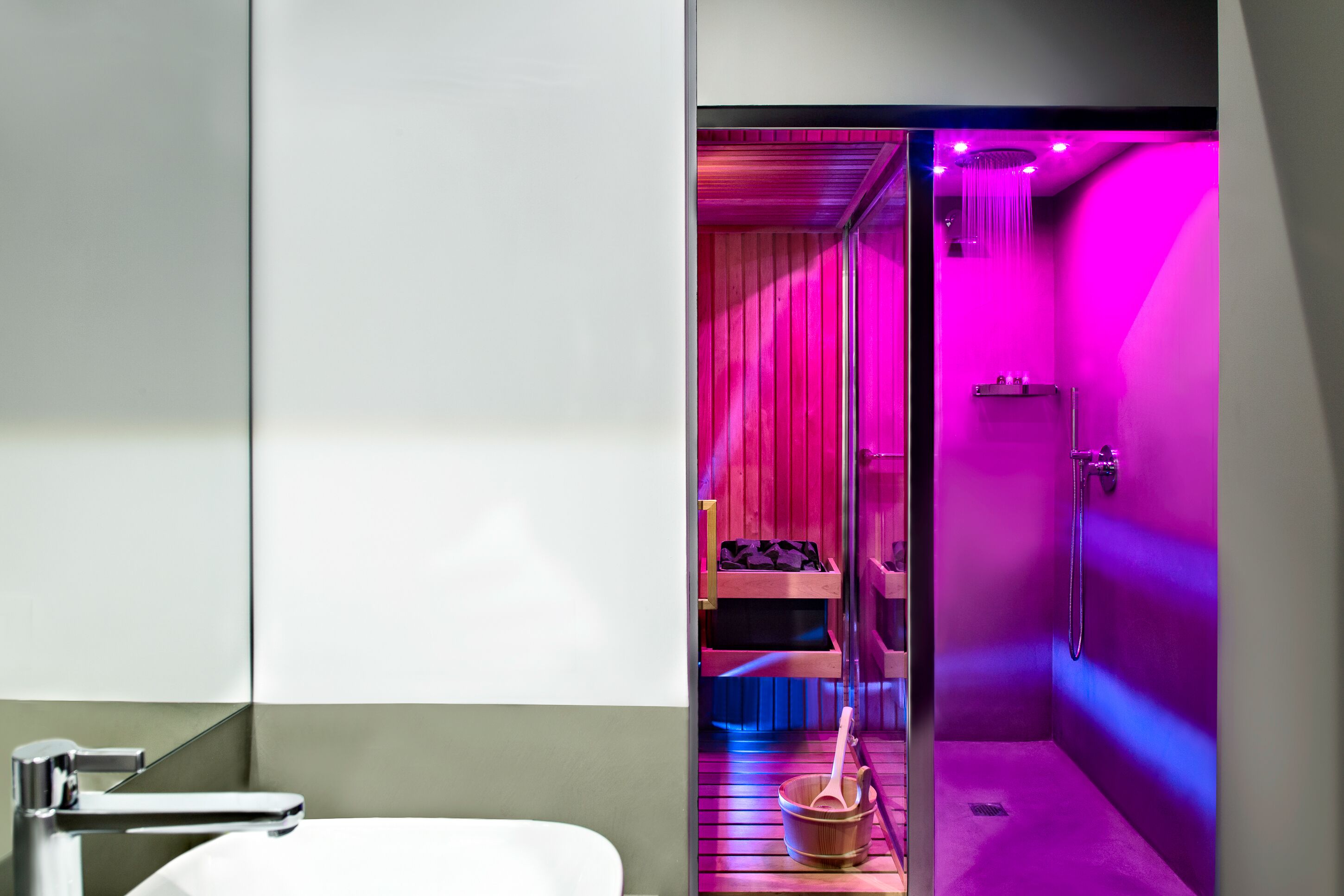 Un bagno moderno con un lavandino bianco, zona doccia vetrata con illuminazione rosa e una panca in legno con un secchio.