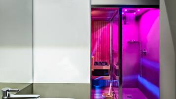 Un bagno moderno con un lavandino bianco, zona doccia vetrata con illuminazione rosa e una panca in legno con un secchio.