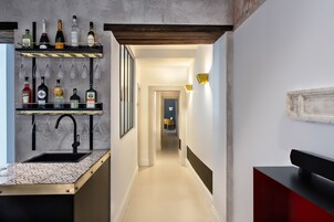 A narrow hallway with a kitchen area on the left, featuring a wine rack, bottles, and a sink. A yellow chair is visible in the distance.