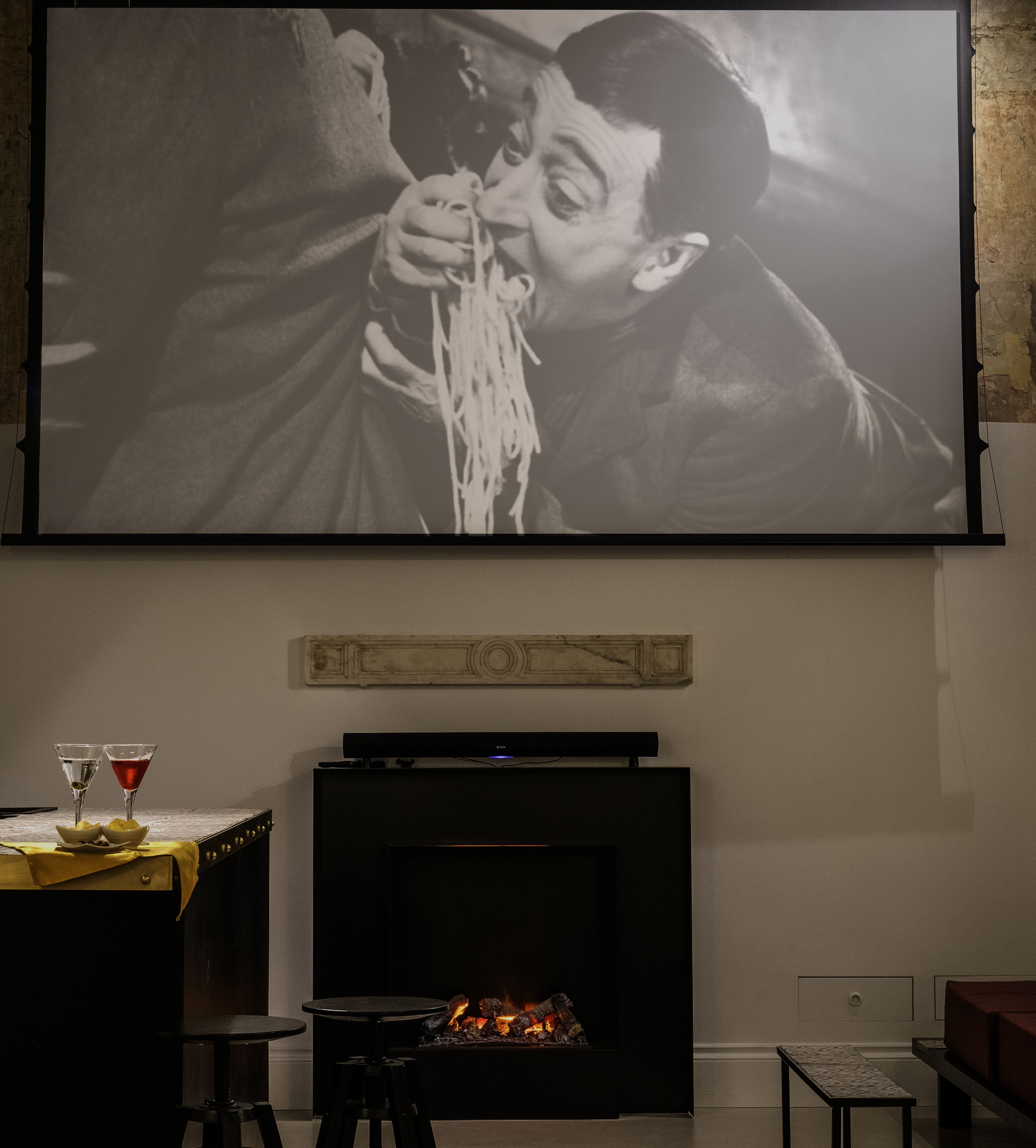 A living room with a flat-screen TV mounted on the wall, a fireplace below, and two bar stools with glasses on a table.