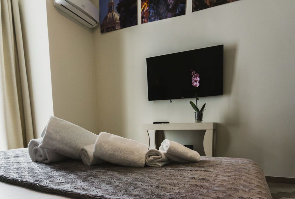 Una cama bien tendida con toallas enrolladas, un televisor de pantalla plana montado en la pared y una mesita con un jarrón de flores.
