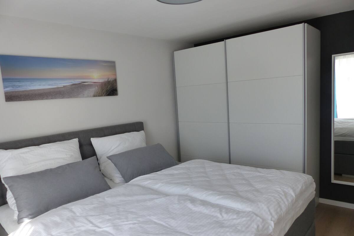 Une chambre à coucher avec un lit, des oreillers gris et blancs, une grande armoire et un tableau représentant une scène de plage accroché au mur.