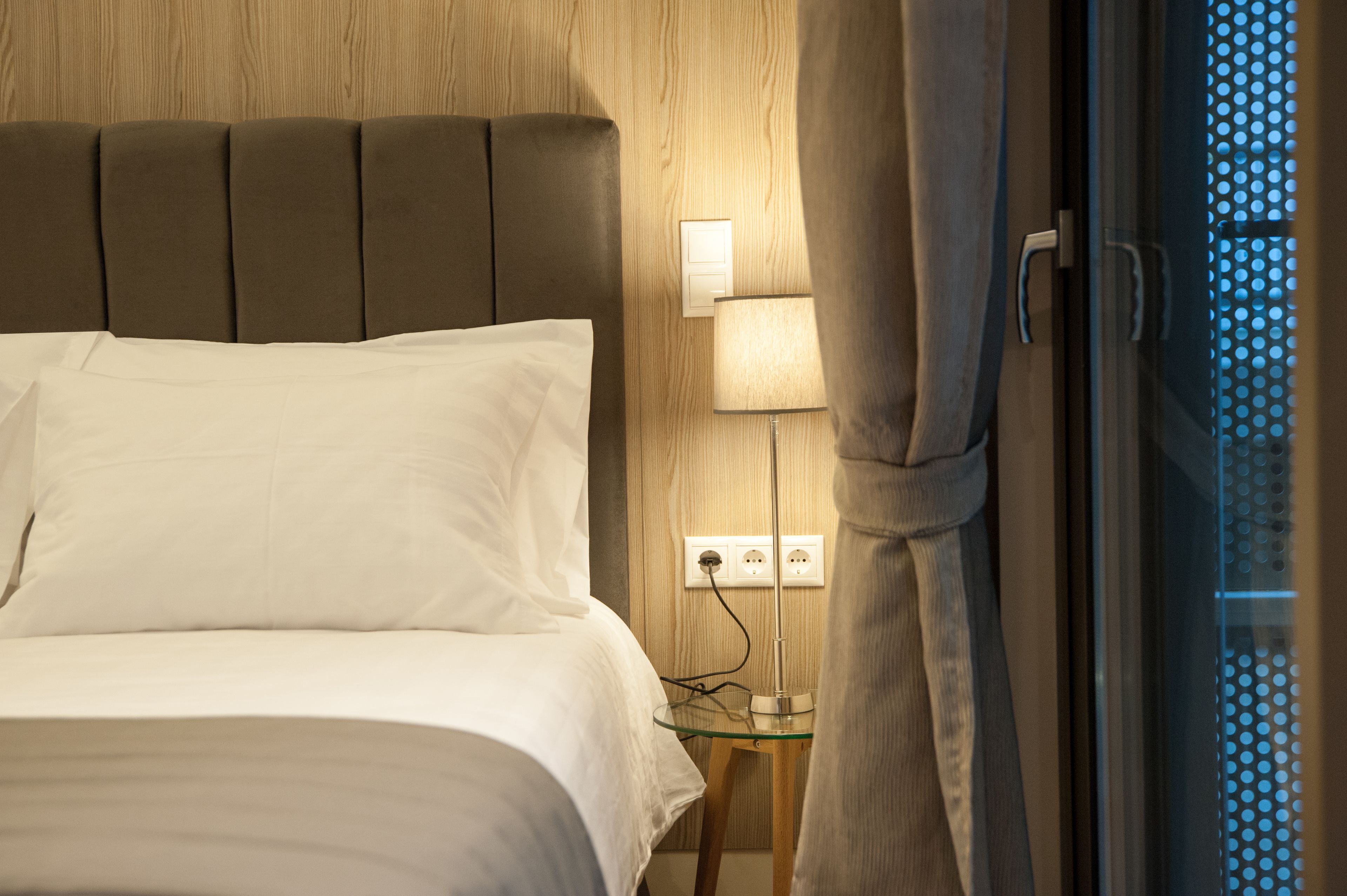Una cama bien hecha con sábanas blancas, una lámpara de pared y un enchufe en una pared de madera.