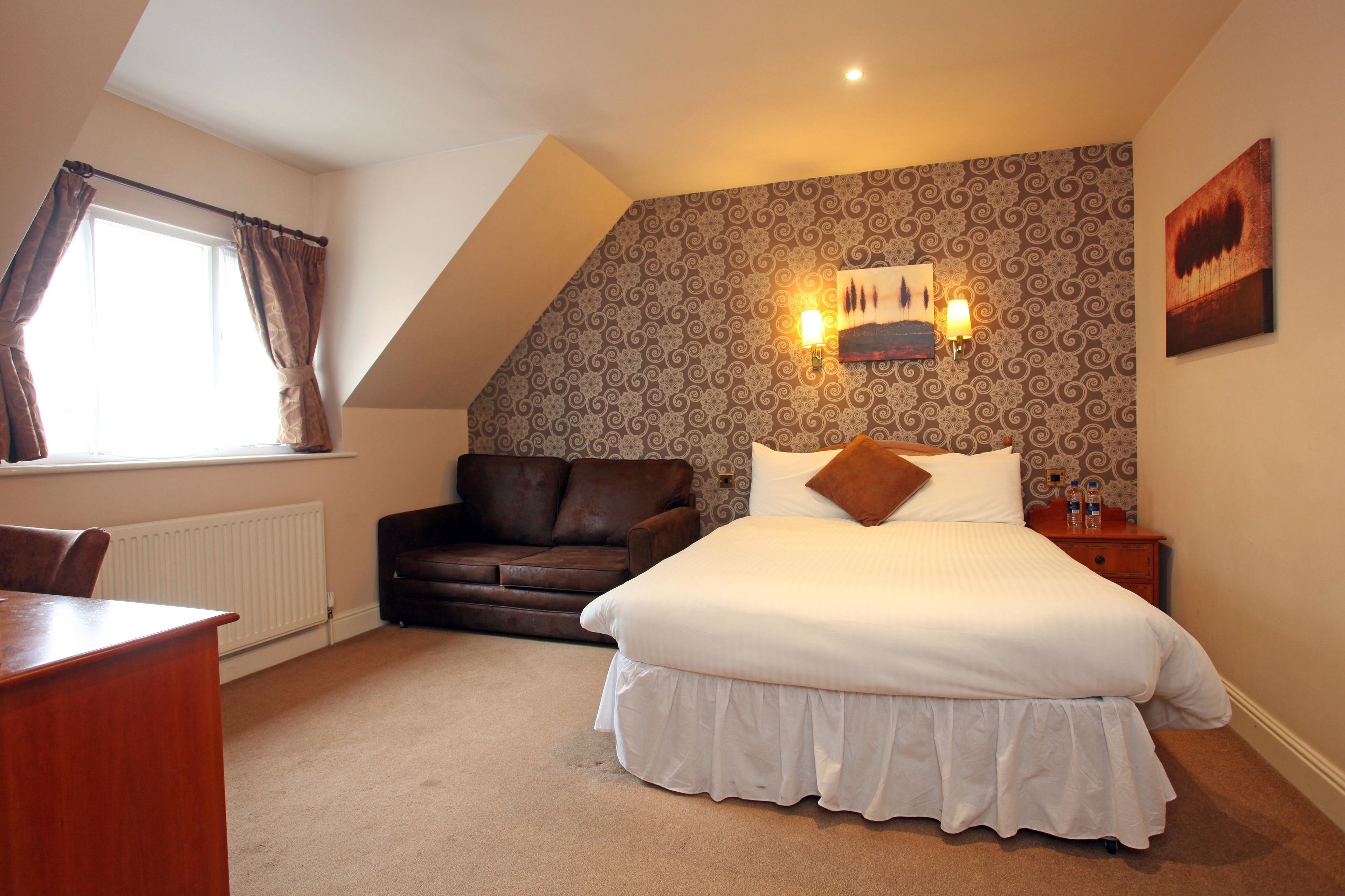 A bedroom with a large bed, a brown leather sofa, a wooden nightstand, and a window with curtains.