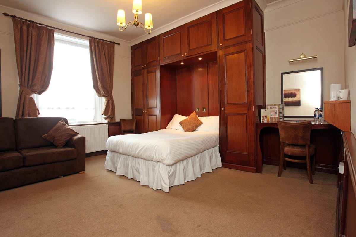 A hotel room with a large bed, a desk, and a chair. There is a window with curtains, a chandelier, and a mirror on the wall.