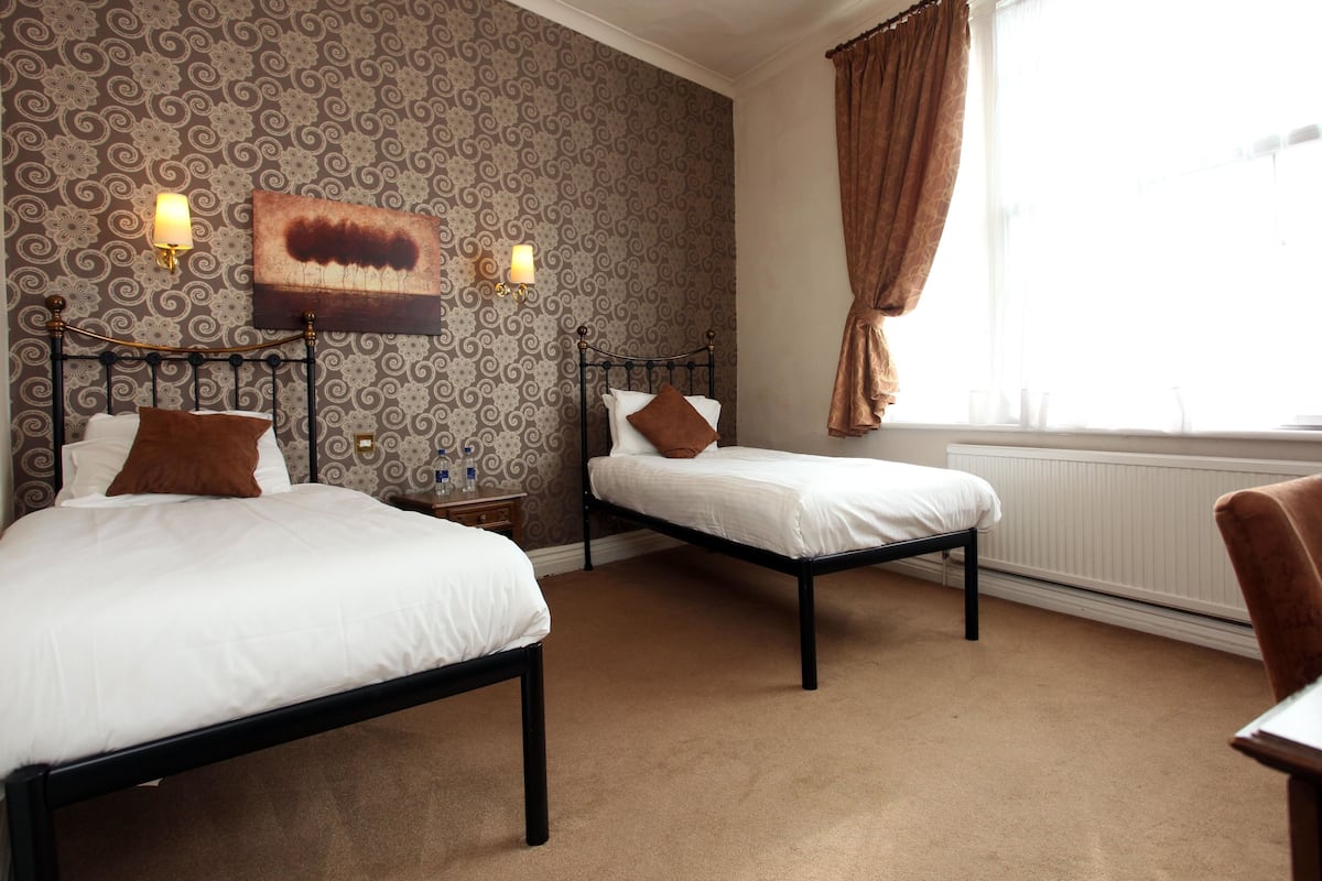A hotel room with two beds, a patterned wallpaper, a painting, and a window with curtains.