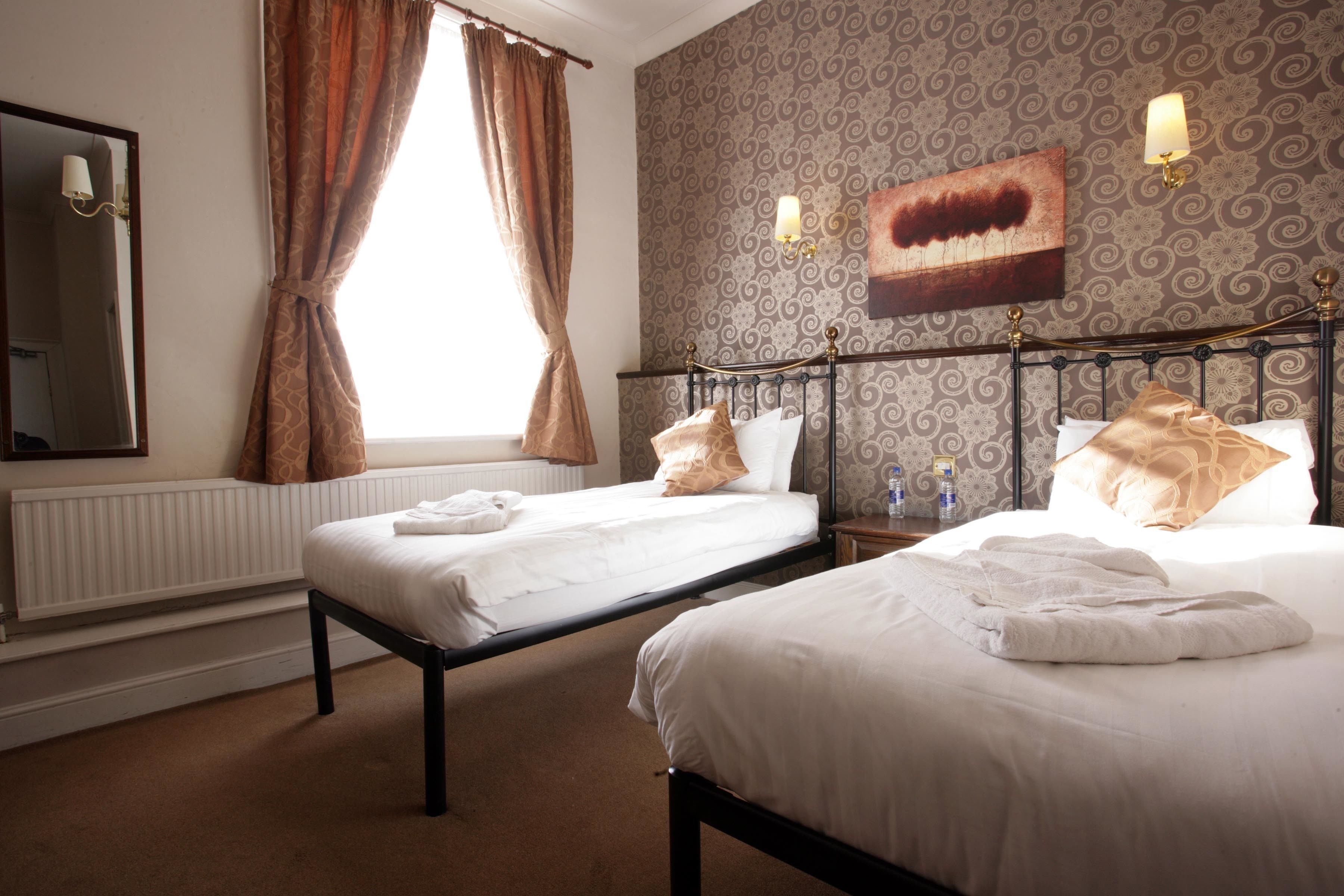 A hotel room with two single beds, a large mirror, a radiator, and patterned wallpaper.