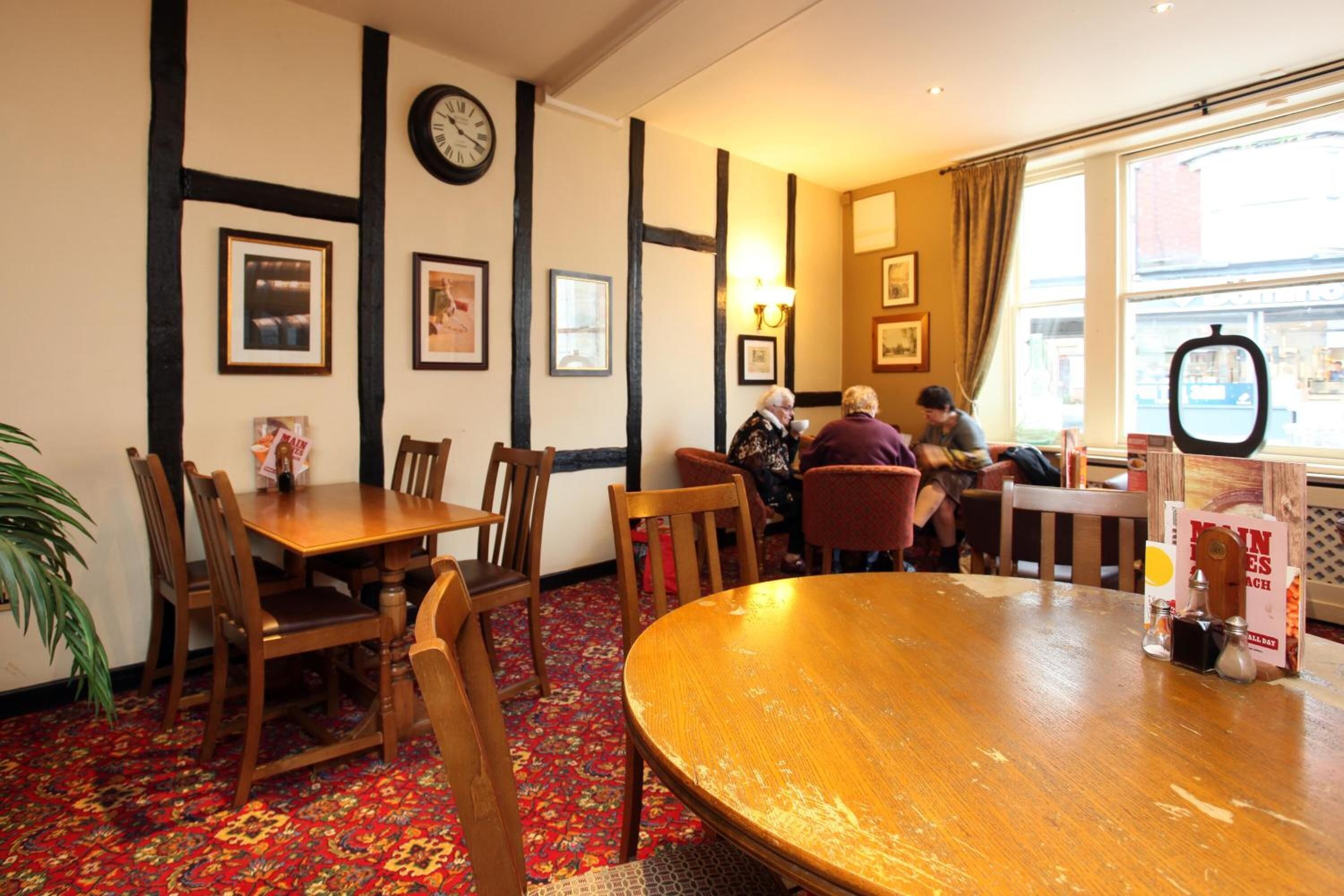 A cozy dining area with wooden tables and chairs, framed pictures on the walls, and a large window with curtains.