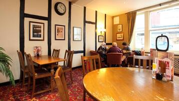A cozy dining area with wooden tables and chairs, framed pictures on the walls, and a large window with curtains.
