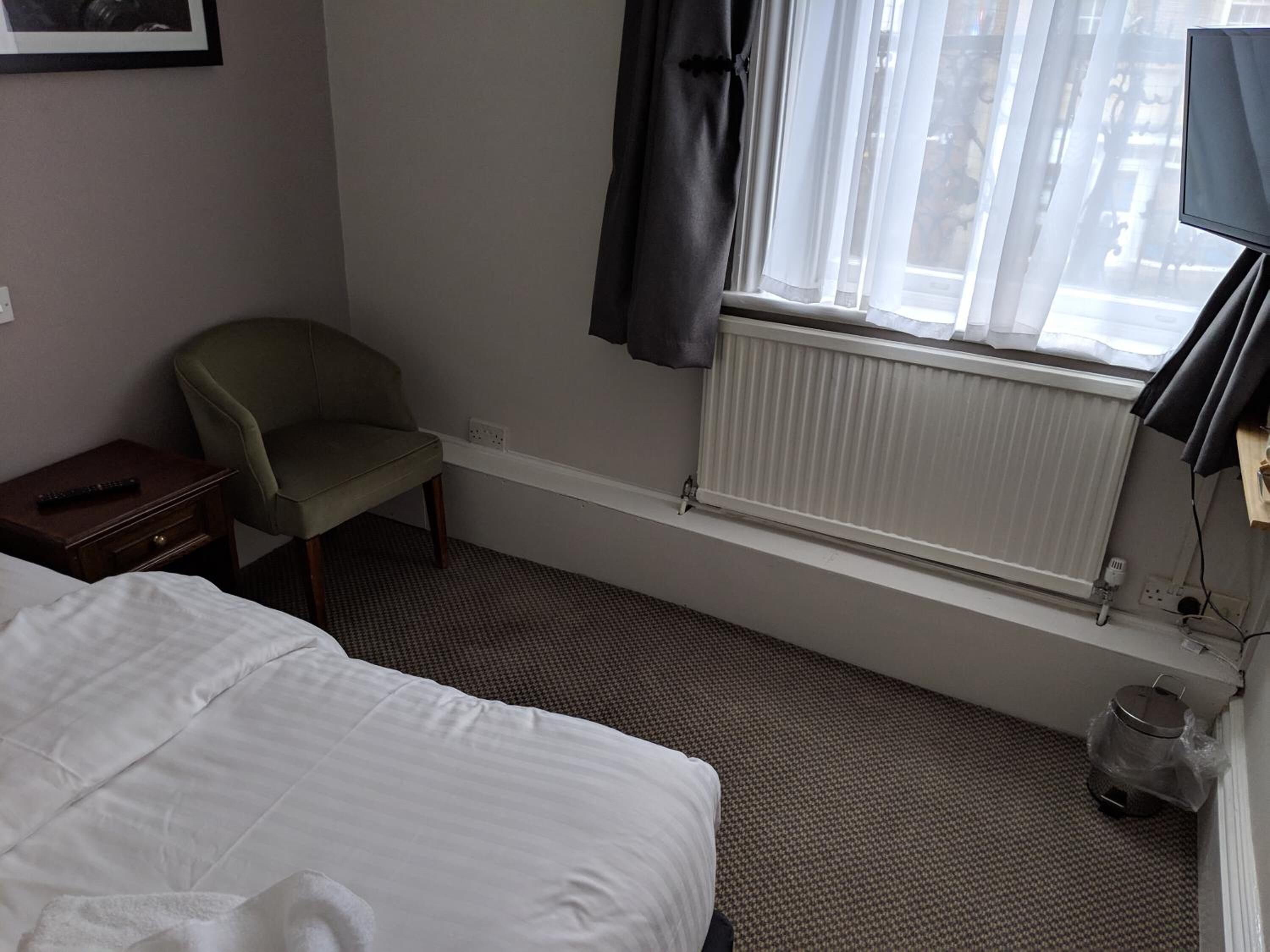 Habitación de hotel con cama, una silla, ventana con cortinas, radiador y un cesto de basura.