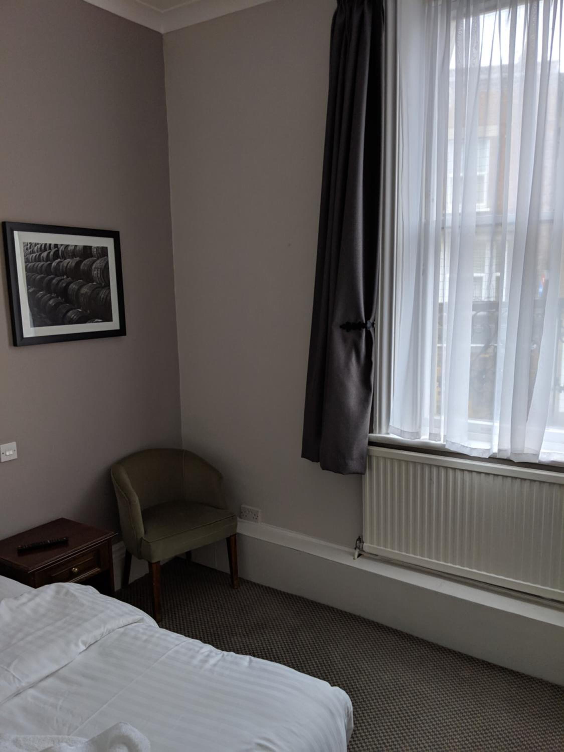 A hotel room with a bed, a chair, a radiator, and a window with curtains.