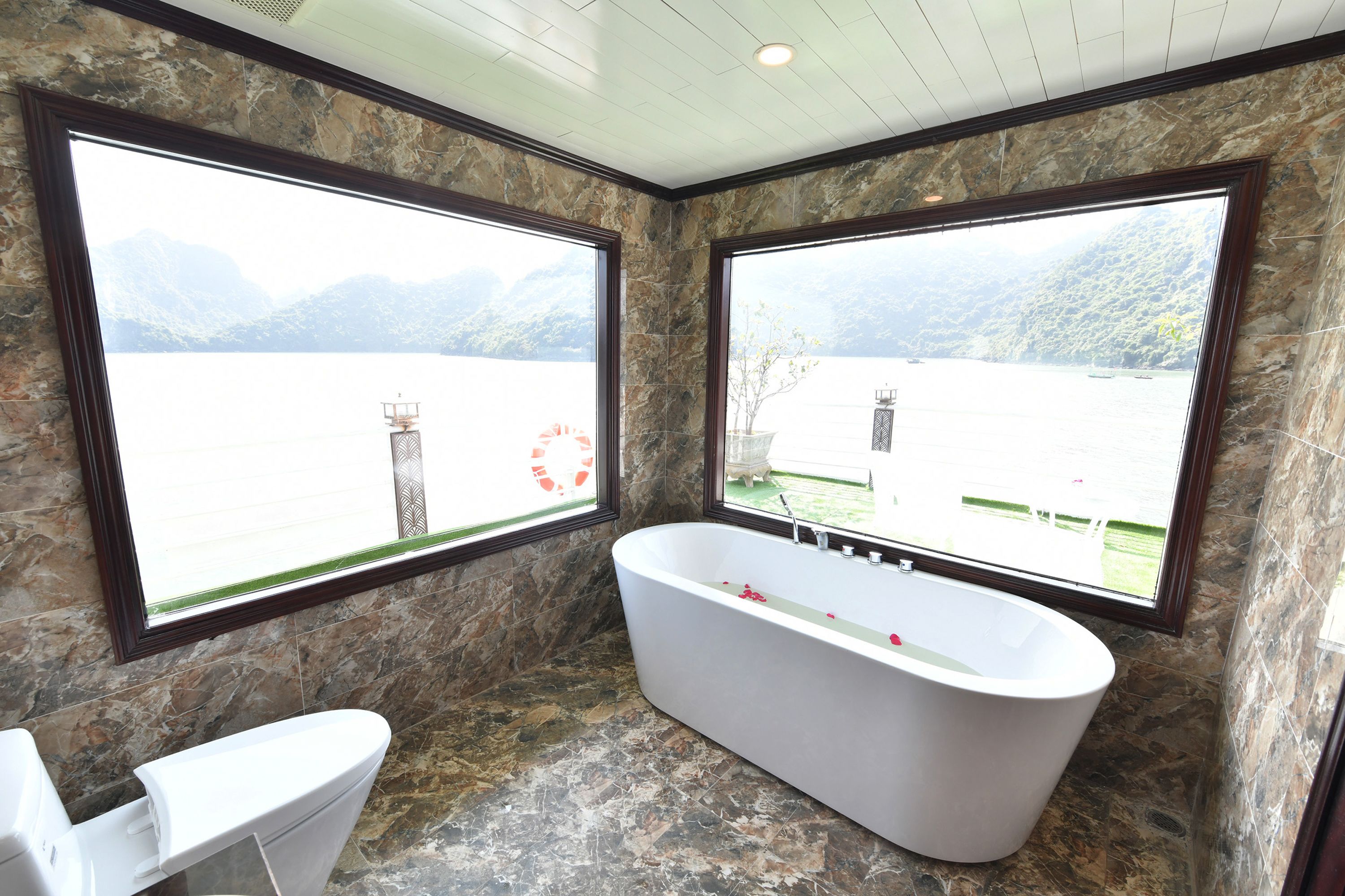 Ein Badezimmer mit freistehender Badewanne, zwei großen Fenstern mit Blick auf die Berge und einer Steinwand.