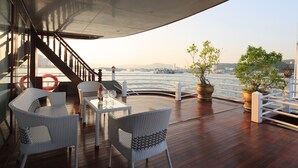 A deck with wicker chairs and a table, overlooking a body of water with boats and a distant cityscape. - Halong Sapphire Cruise (Tuan Chau)