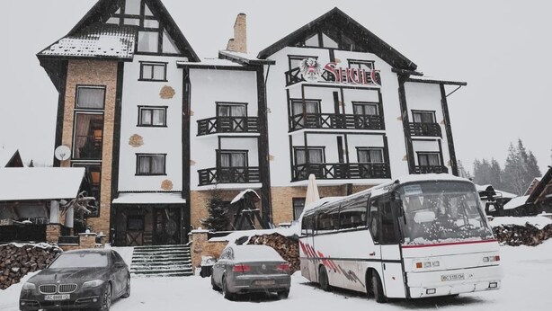 A multi-story building with a ski resort sign, a bus, and cars parked in front. - Hotel Shulc (Bukovel)