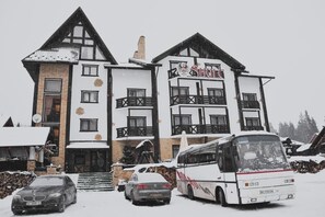 A multi-story building with a ski resort sign, a bus, and cars parked in front. - Hotel Shulc (Bukovel)