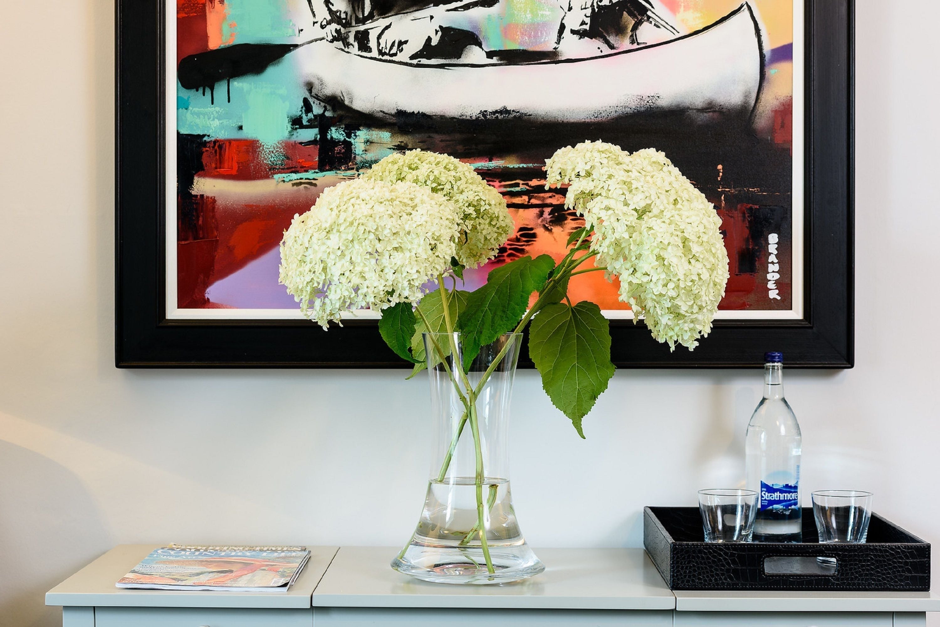Un florero de vidrio transparente con flores blancas sobre una mesa blanca, un cuadro abstracto enmarcado arriba y una botella con vasos sobre una bandeja negra.