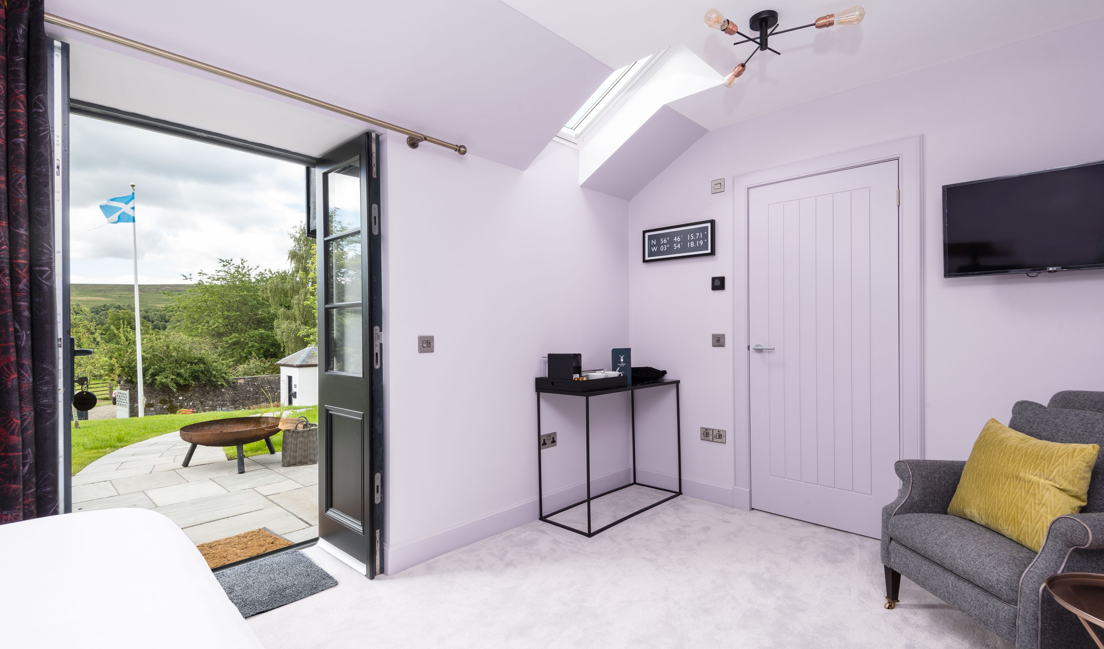 A modern room with a large sliding glass door leading to a garden, a flat-screen TV mounted on the wall, and a cozy seating area with a grey sofa and a yellow pillow.