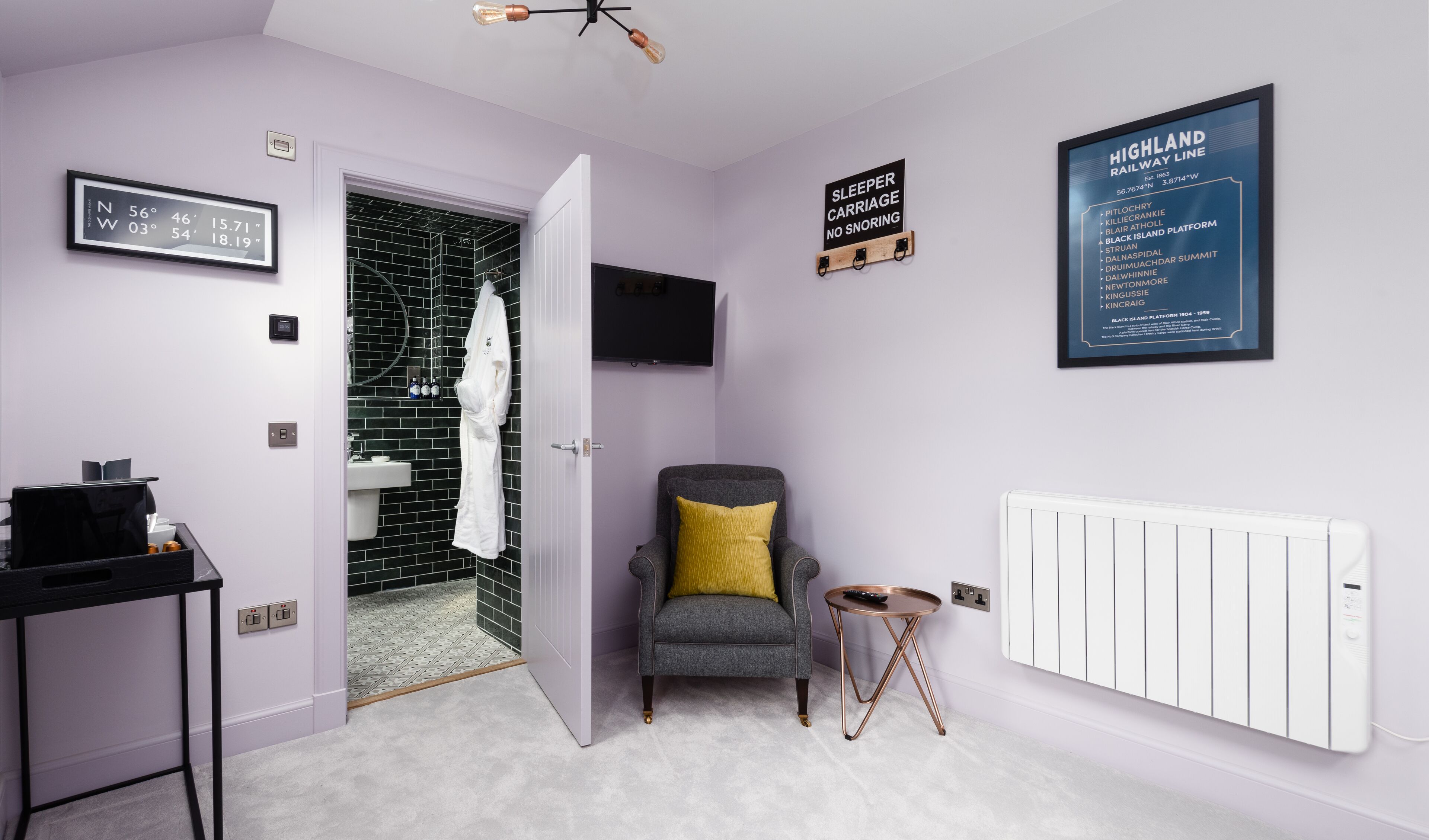 A modern bathroom with a black and white tiled shower, a white sink, and a grey armchair with a yellow pillow. There are framed maps and a sign on the wall.