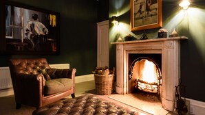 A living room with a fireplace, a leather sofa, a cushioned ottoman, a painting on the wall, and a lamp. - The Old Manse of Blair (Pitlochry)