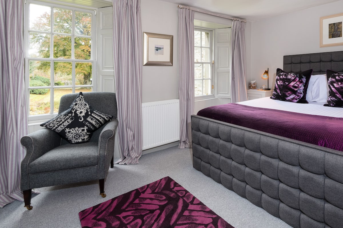 A bedroom with a grey upholstered armchair, a large bed with a grey upholstered headboard, and a purple throw blanket.