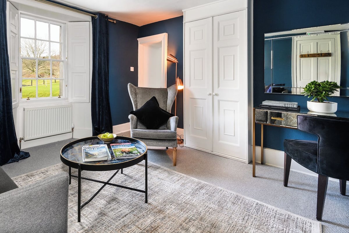 A room with a large window, a chair, a table with magazines, a desk with a computer, and a mirror.