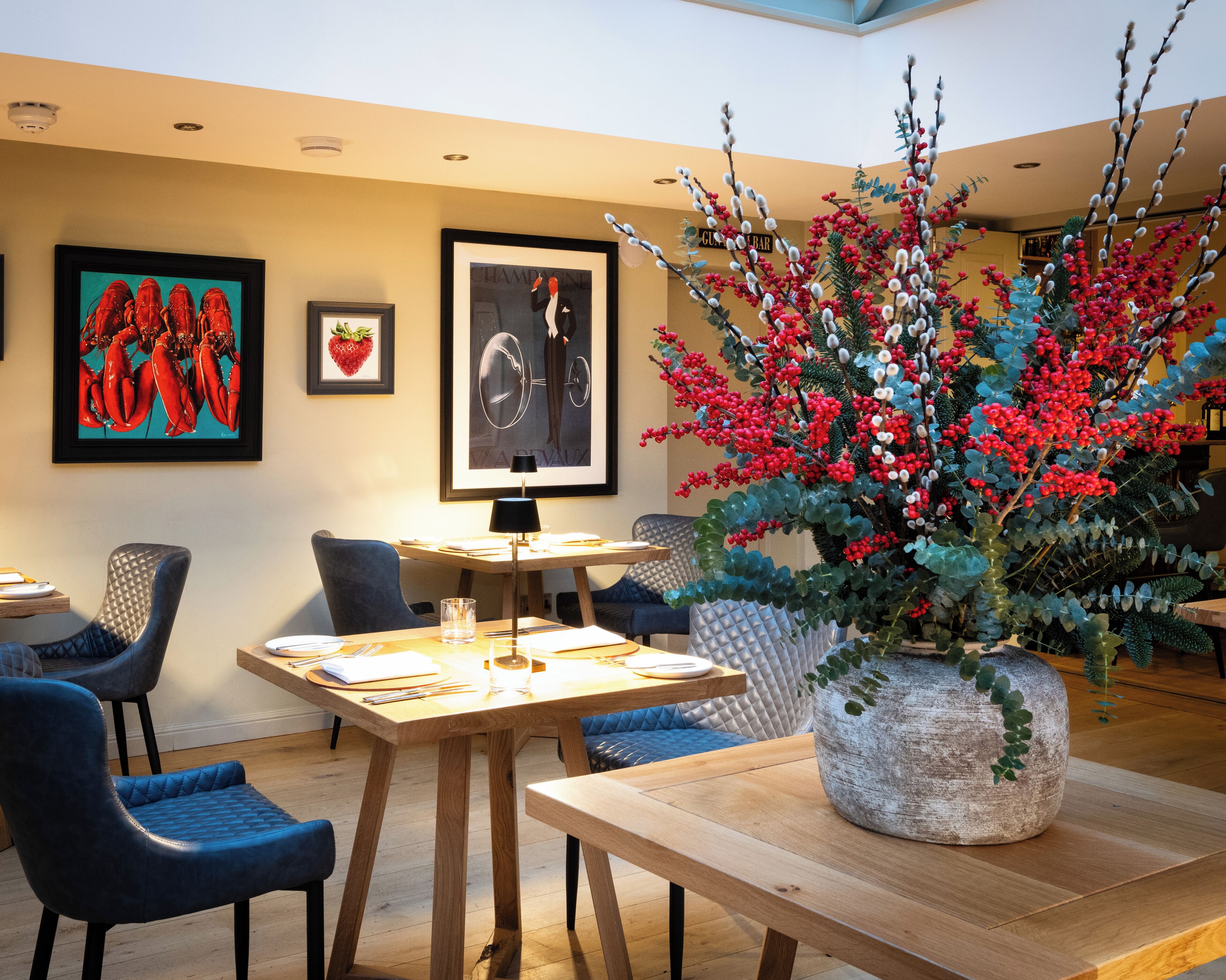 A dining area with a wooden table, blue upholstered chairs, and a large decorative plant with red berries.