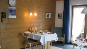 A cozy room with a wooden wall, a table set with breakfast items, a clock, and a chair. - Ferienapartmenthaus Hubertushof (Mittenwald)