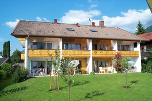 A two-story building with a tiled roof, balconies with wooden railings, and a well-maintained lawn with small trees. - Ferienapartmenthaus Hubertushof (Mittenwald)