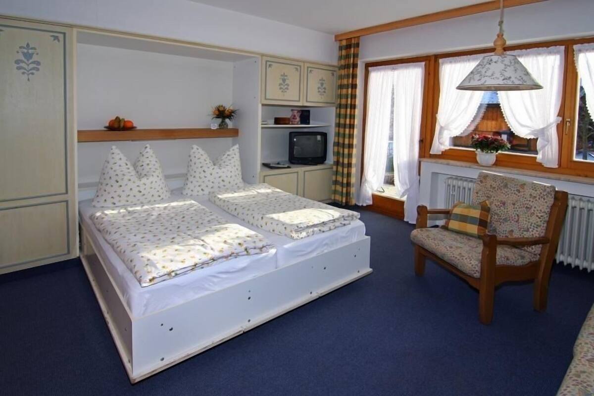 A bedroom with a double bed, a chair, a television, and a window with curtains.