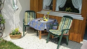 A small patio with a table and two chairs, a wicker basket with flowers, and a window with curtains. - Ferienapartmenthaus Hubertushof (Mittenwald)