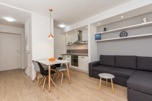 A modern living room with a grey sofa, a dining table with chairs, a small round table, and a kitchen area with cabinets and a stove. - Bell Apartment (Zadar)