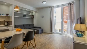Un salon moderne avec un canapé, une table à manger et des chaises. On y trouve également un coin cuisine avec des placards et une machine à café. Une grande fenêtre avec des rideaux est visible.