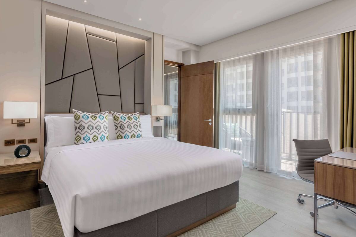 A modern hotel room with a large bed, a desk, and a view of the city through sheer curtains.