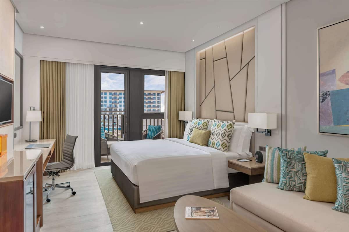 A hotel room with a bed, a desk, a chair, a sofa, and a large window offering a view of buildings.