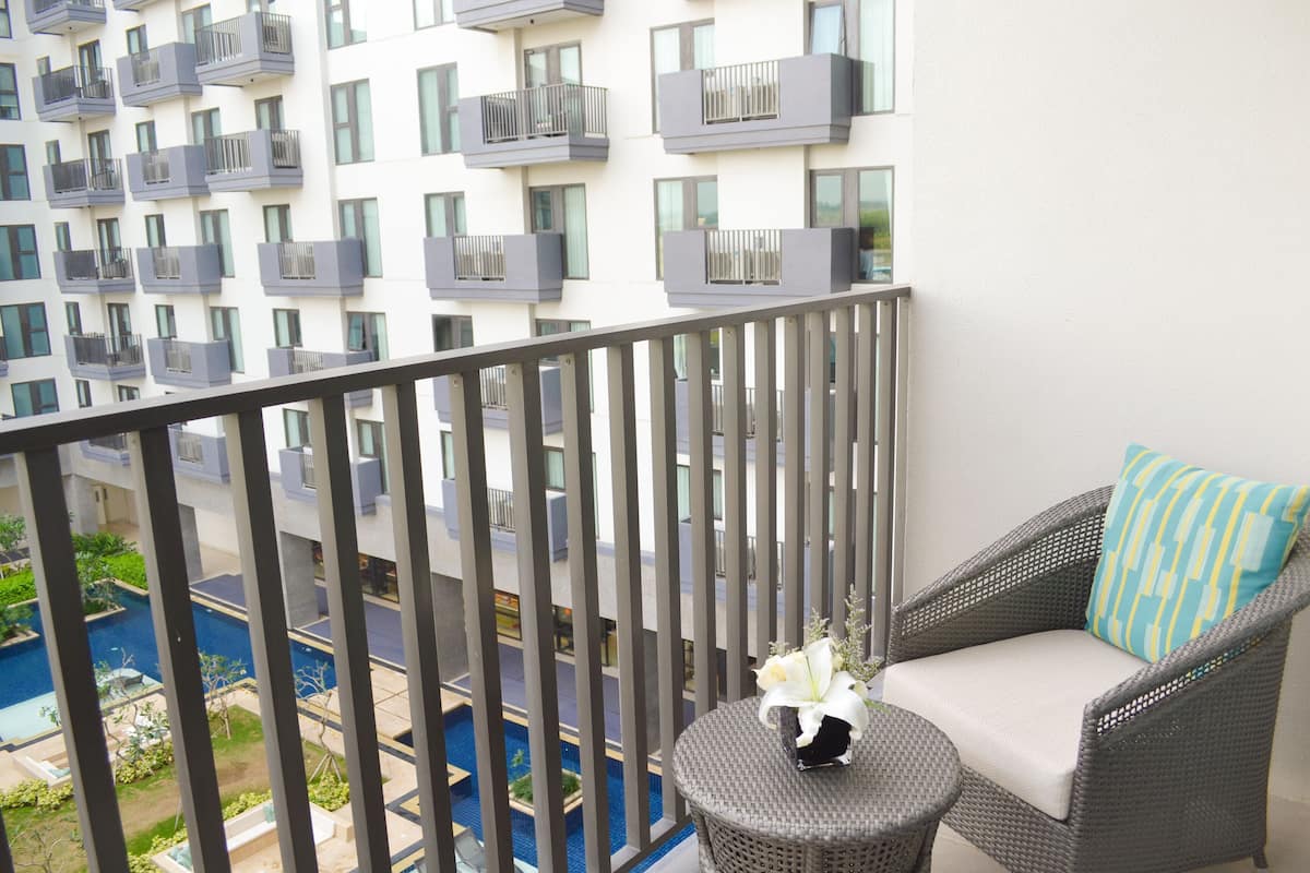 A balcony with a wicker chair and a small round table, overlooking a swimming pool and a building with balconies.