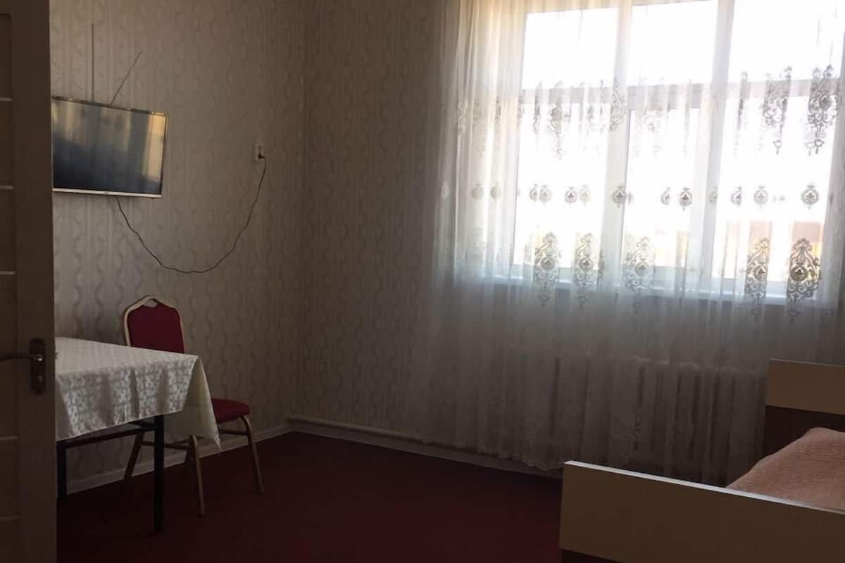 A room with a bed, a table with a chair, a TV mounted on the wall, and a window with curtains.