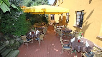 An outdoor patio with wicker chairs and tables, covered by a yellow umbrella, and surrounded by greenery.