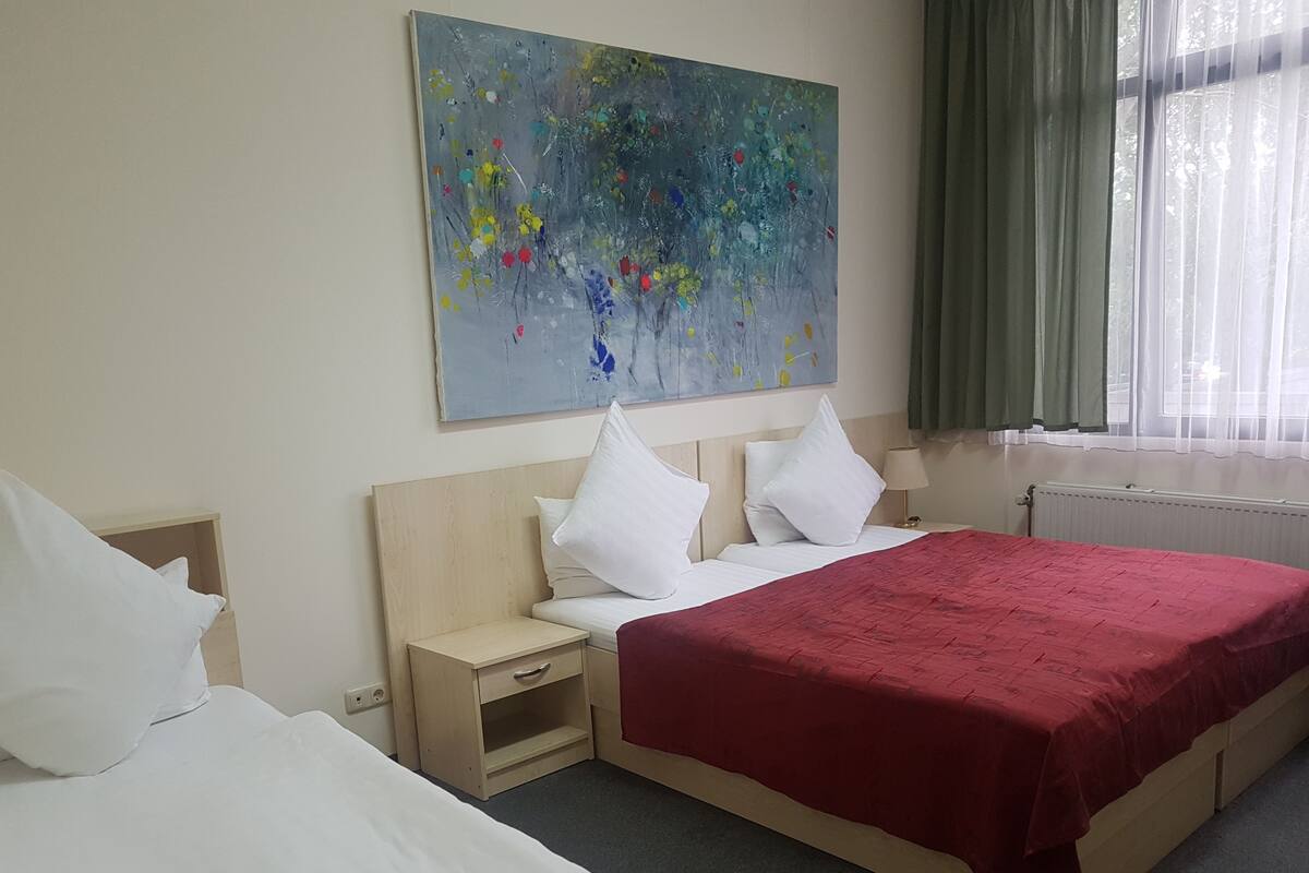 Une chambre d’hôtel avec un grand tableau abstrait, un lit recouvert d’une couverture rouge, une table de chevet et une fenêtre avec des rideaux.