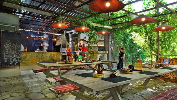 An outdoor restaurant with wooden tables and benches, a bar area, and a chef preparing food. - Berlin Art Hotel (Gyumri)