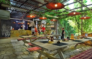 An outdoor restaurant with wooden tables and benches, a bar area, and a chef preparing food. - Berlin Art Hotel (Gyumri)