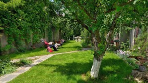 A garden with a stone pathway, a tree, and two people sitting on chairs reading. - Berlin Art Hotel (Gyumri)
