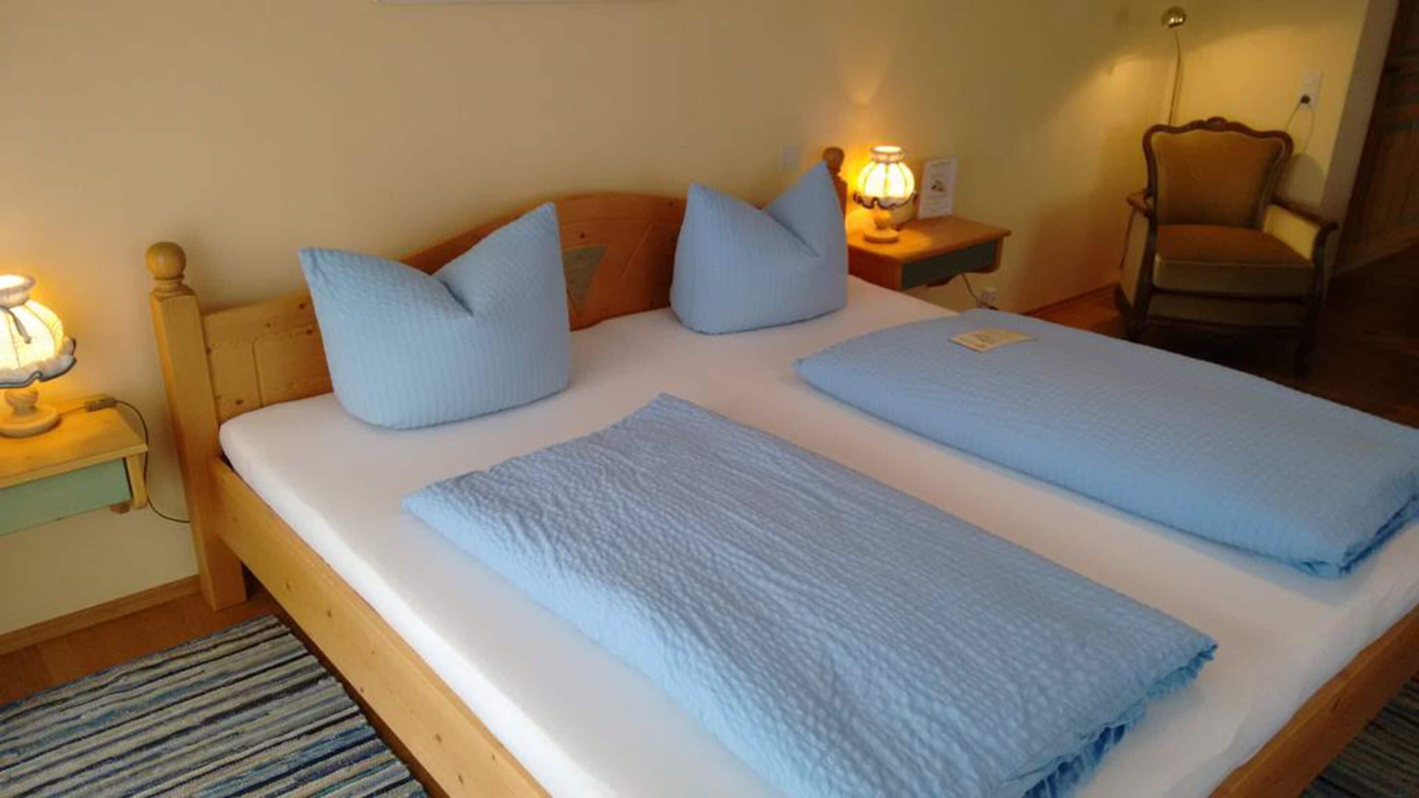 A double bed with two pillows and a blanket, bedside tables with lamps, a chair, and a wooden headboard.
