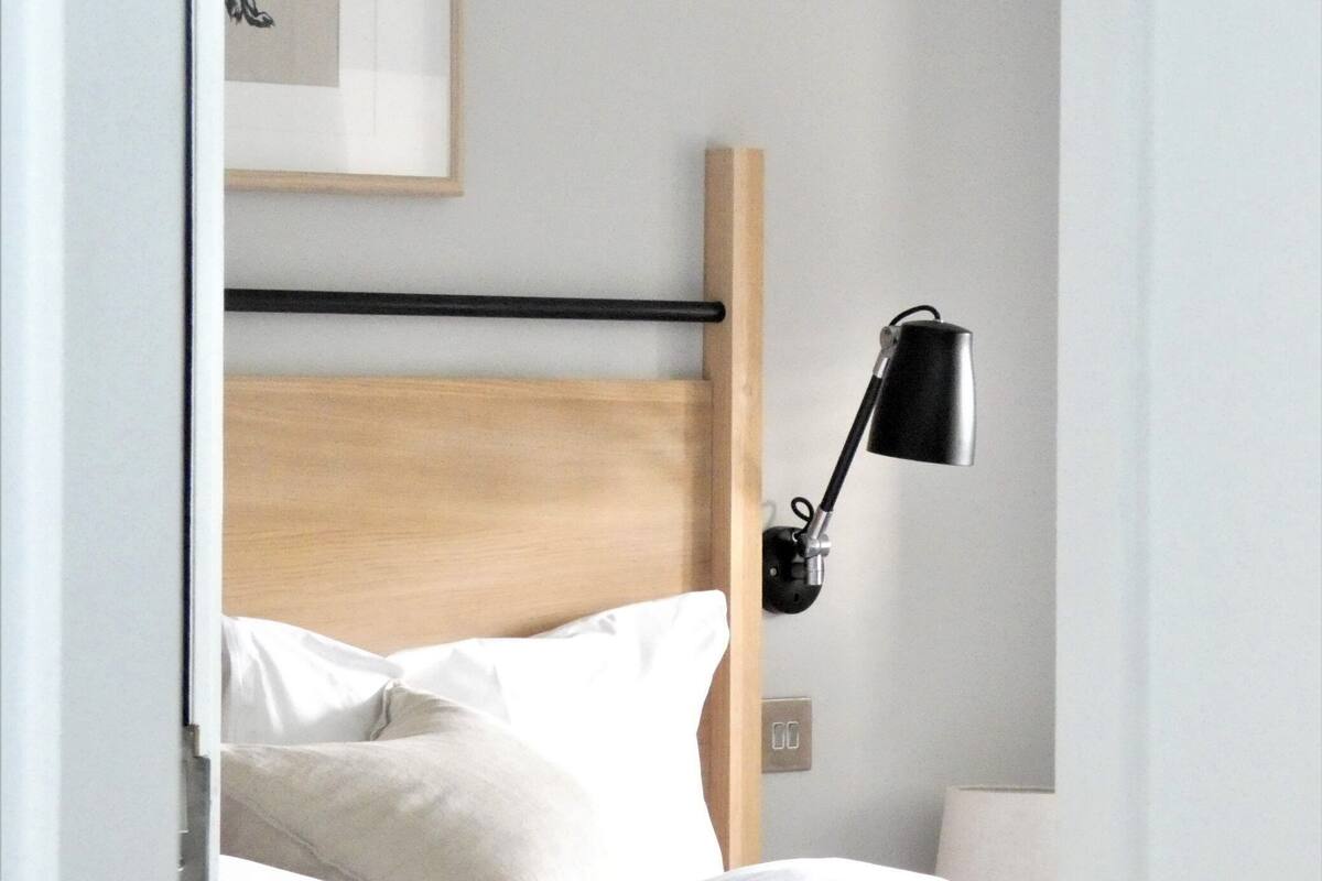 Cama bem arrumada com roupa de cama branca, cabeceira de madeira, abajur preto e quadro pendurado na parede.