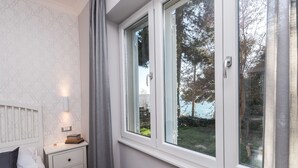 A bedroom with a bed, bedside table, lamp, and a window with a view of trees and a body of water. - Sunset Beach Apartment (Zadar)
