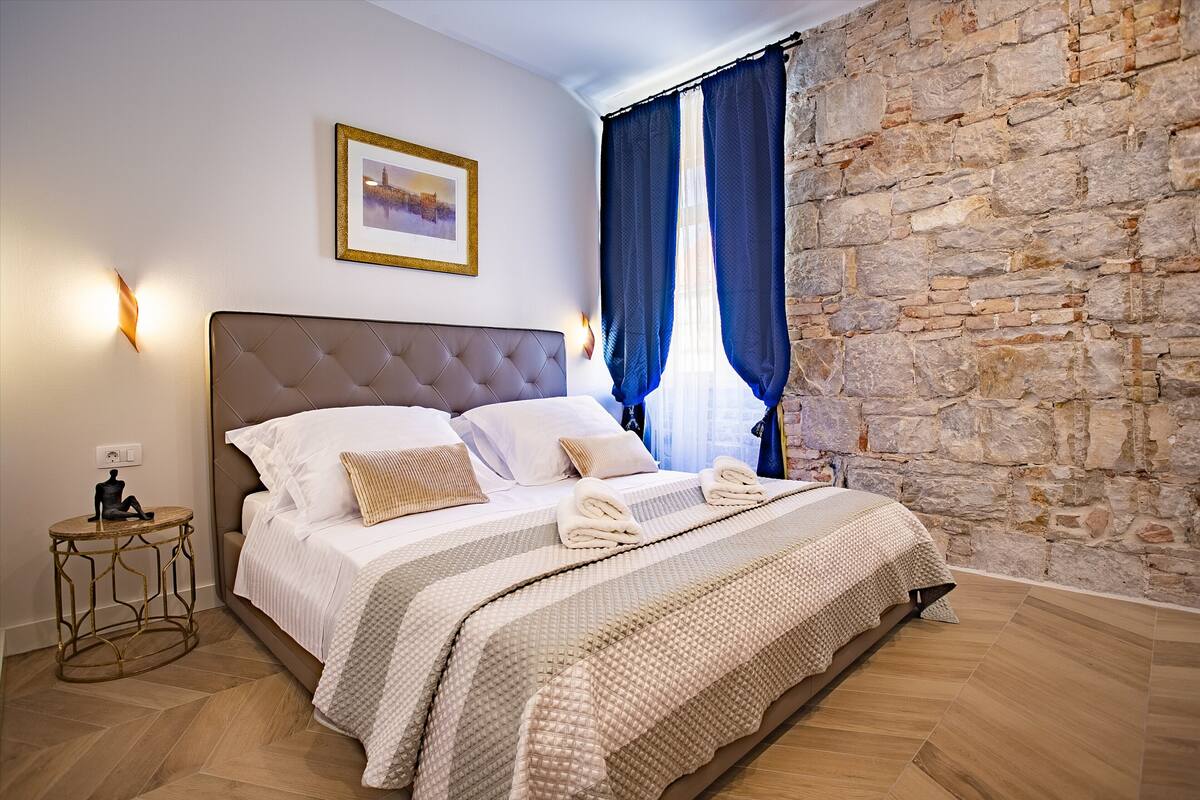 A bedroom with a stone wall, a bed with a tufted headboard, and a small round table with a lamp.